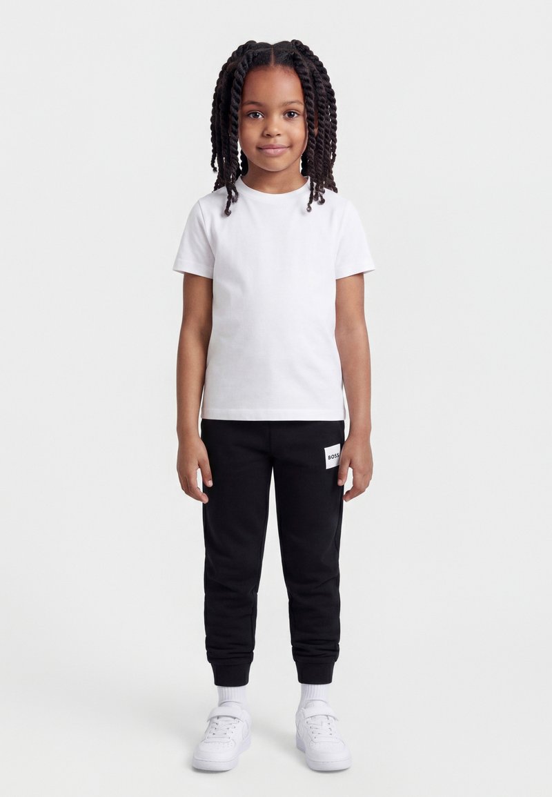 Κορίτσι με πλεξούδες που στέκεται, φοράει ένα απλό λευκό μπλουζάκι, μαύρο παντελόνι φόρμας με λογότυπο, λευκές κάλτσες και λευκά αθλητικά παπούτσια.