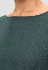 Top verde azulado con textura acanalada, escote ancho y acabado suave. La tela parece ligera y transpirable con un brillo sutil.