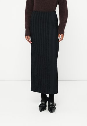 Jupe maxi noire à côtes avec un motif texturé, taille haute et légère volute en bas. Associée à des chaussures noires à bout pointu.