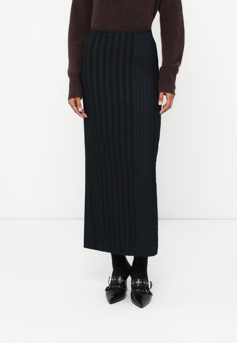 Jupe maxi noire à côtes avec un motif texturé, taille haute et légère volute en bas. Associée à des chaussures noires à bout pointu.