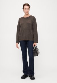 Brun strikket genser med ribbestrikket kant og knapper på siden. Sammen med mørke, utsvingte jeans og svarte slip-on sko. Holder en liten svart håndveske.