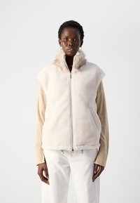 Gilet in pelliccia sintetica di un beige chiaro con colletto alto, chiusura a zip e tasche laterali sopra una camicia beige a maniche lunghe. Pantaloni bianchi a gamba larga.
