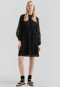 Vestido negro con mangas largas transparentes, tejido texturizado, cintura ajustada y cinturón a juego. Combinado con zapatos estampados y calcetines negros.