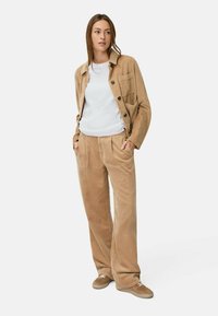 Corduroy jasje en broekset in lichtbruin, met een knoopsluiting, voorzak en een ontspannen pasvorm. Gecombineerd met een witte t-shirt en sneakers.