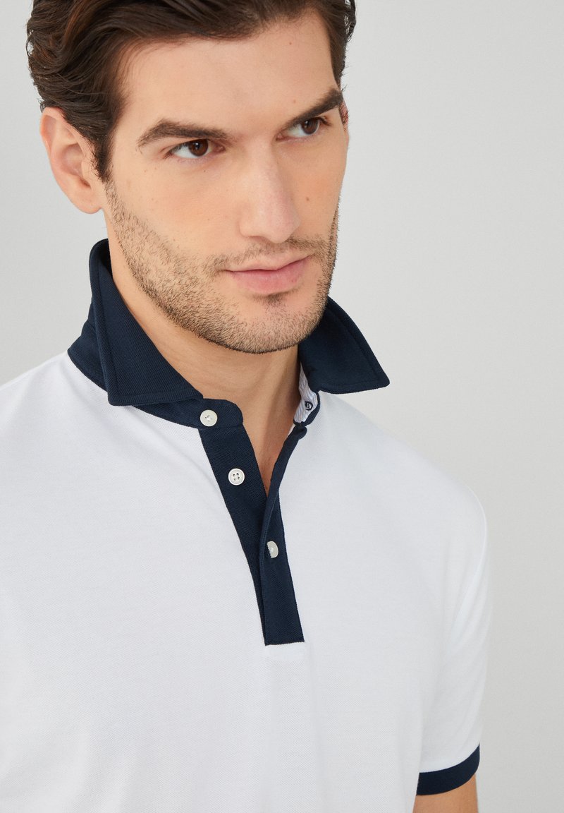 Polo blanc avec un col marine et des boutons accentués. Présente un tissu texturé et une coupe classique.