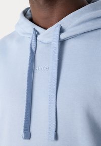 Gros plan d'un sweat à capuche bleu clair avec des cordons épais et "HUGO" embossé en bleu assorti sur la poitrine.