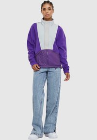 Lilla og grå jakke med høy krage og glidelås; har fleece-tekstur med oversized passform og kontrastpaneler.
