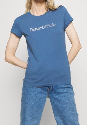 Frau in einem blauen Marc O’Polo T-Shirt und blauer Jeans, stehend mit den Händen hinter dem Rücken vor einem schlichten Hintergrund.