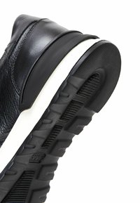 Gros plan sur une basket en cuir noir montrant une semelle noire texturée avec un profond profil et une bande intercalaire blanche sur un fond blanc.
