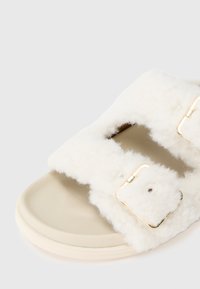 Des sandales à glissière blanches bordées de fourrure, avec deux bretelles réglables et des détails en métal doré. La semelle est beige avec une texture lisse.