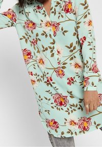 Tunique fleurie bleu clair, avec motifs floraux rouges et blancs, manches longues et fermeture boutonnée sur le devant. Texture de tissu douce.