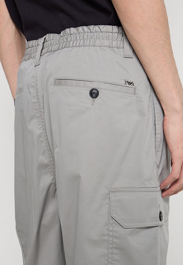 Pantalones cargo gris claro con cintura elástica, bolsillos laterales y detalles de botones. Presentan una textura suave y un diseño minimalista.