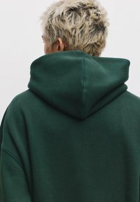 Felpa con cappuccio verde scuro e grande cappuccio arrotondato. Tessuto morbido e testurizzato con cuciture visibili lungo i bordi. Design semplice e minimale.