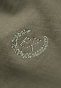 Tessuto oliva con logo ricamato contenente le iniziali "E" e "X" racchiuse in una corona di alloro, in una tonalità di verde più chiara.