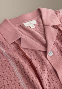 Ružový pletený kardigán s otvoreným vzorom, golierovým výstrihom, zapínaním na gombíky vpredu a pruhovanými detailmi v tmavších a svetlejších ružových odtieňoch.