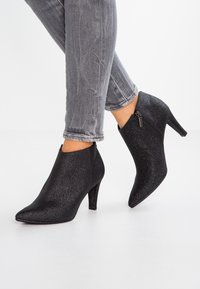 Stivaletti neri con glitter, punta affusolata, design elegante e chiusura con zip laterale. Ambientati su jeans skinny grigi.