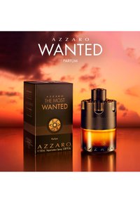 Azzaro The Most Wanted parfymeflaske formet som en revolversylinder ved siden av sin svarte og gullfargede eske, mot en varm oransje solnedgang.