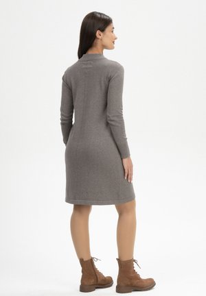 Vrouw met lang donker haar, gekleed in een grijze, knielange jurk met lange mouwen en bruine veterlaarzen, staand met een lichte draai van het lichaam.