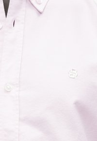 Chemise boutonnée violet clair en tissu texturé. Présente des boutons blancs et un petit logo brodé sur le côté gauche.