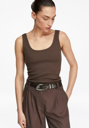 Kobieta z ciemnymi włosami związanymi do tyłu, ubrana w brązowy prążkowany bezrękawnik, pasujące brązowe spodnie oraz czarny pasek ze srebrną klamrą.