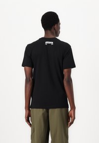 Black cotton t-shirt with short sleeves, featuring a small white logo at the back neckline. Worn with olive green cargo pants.