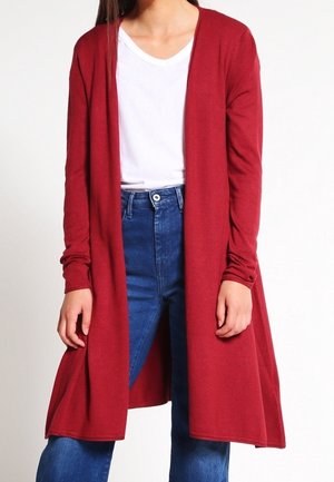 Femme portant un long cardigan rouge ouvert sur un t-shirt blanc et un jean bleu taille haute, debout devant un fond uni.