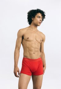 Sarkans kokvilnas bokseršortus ar Calvin Klein logo jostas daļu. Dizains ietver pieguļošu formu un gludu tekstūru. Modelis rāda sānu profilu.