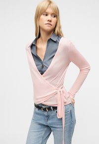 Γυναίκα με ξανθά μαλλιά που φοράει γκρι πουκάμισο με γιακά κάτω από ένα ροζ ανοιχτόπλευρο πουλόβερ, ανοιχτό μπλε τζιν και μαύρη ζώνη, με φόντο λευκό.