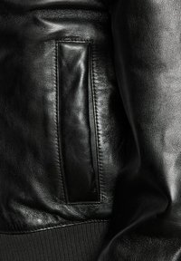Schwarze Lederjacke mit glatter Textur, seitlichen Taschen und gerippten Bündchen. Der Stoff ist elegant mit subtiler glänzender Oberflächenstruktur.