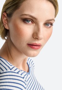 Portret osobe s kratkom plavom kosom, koja nosi prugastu plavu i bijelu majicu. Glatka koža, lagani make-up i plave oči.