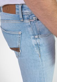 Los jeans de mezclilla de color azul claro presentan una textura suave, un diseño de cinco bolsillos, un parche de cuero marrón y herrajes plateados en los bolsillos.