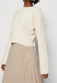 Frau trägt einen cremefarbenen, gerippten Strickpullover, kombiniert mit einem glitzernden, gefalteten beigen Rock vor einem schlichten Hintergrund.