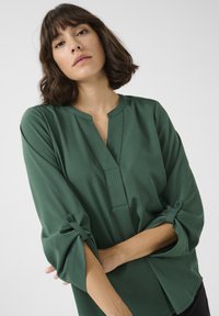Blusa verde a maniche lunghe con scollatura a V e polsini con bottoni. Tessuto liscia con una texture sottile e vestibilità ampia.