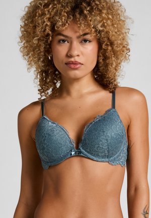 Vrouw met krullend haar die een blauwe kanten push-up bh draagt met dunne bandjes en een klein strikje in het midden, staand tegen een effen achtergrond.