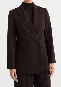 Hnědý dvouřadý blazer, přiléhavý střih, vyrobený z hladkého materiálu. Má vyštípnuté klopy, dvě přední kapsy a černé knoflíky.
