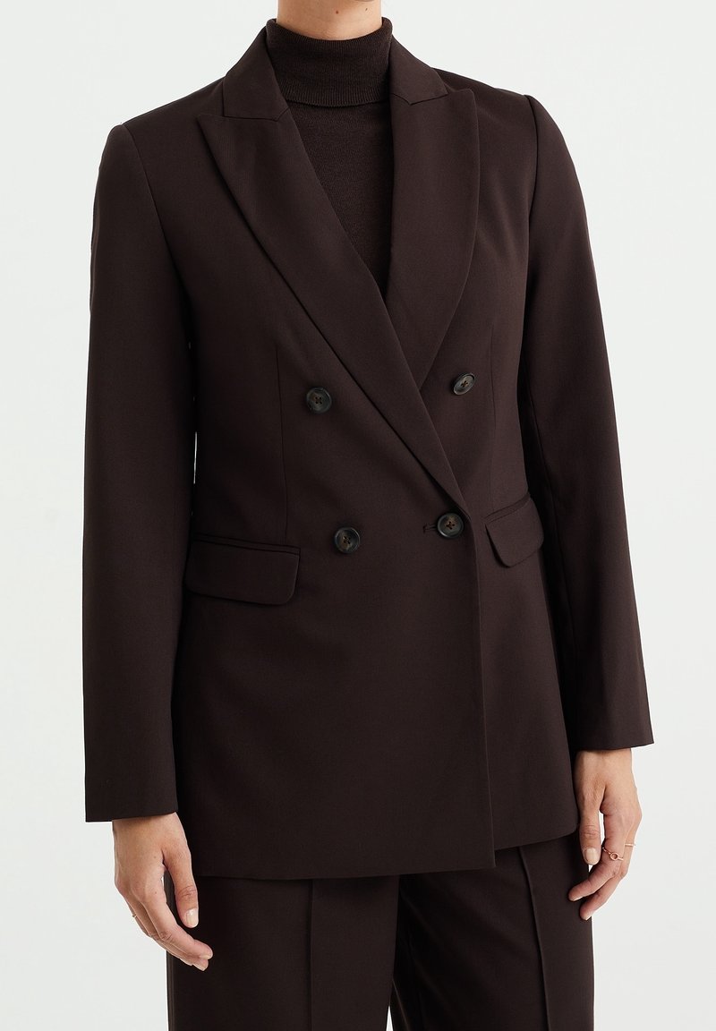 Hnědý dvouřadý blazer, přiléhavý střih, vyrobený z hladkého materiálu. Má vyštípnuté klopy, dvě přední kapsy a černé knoflíky.
