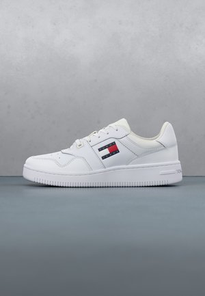 Białe niskie trampki z zapięciem na sznurowadła, z logo Tommy Jeans po boku oraz teksturowaną podeszwą na szarej powierzchni i tle.