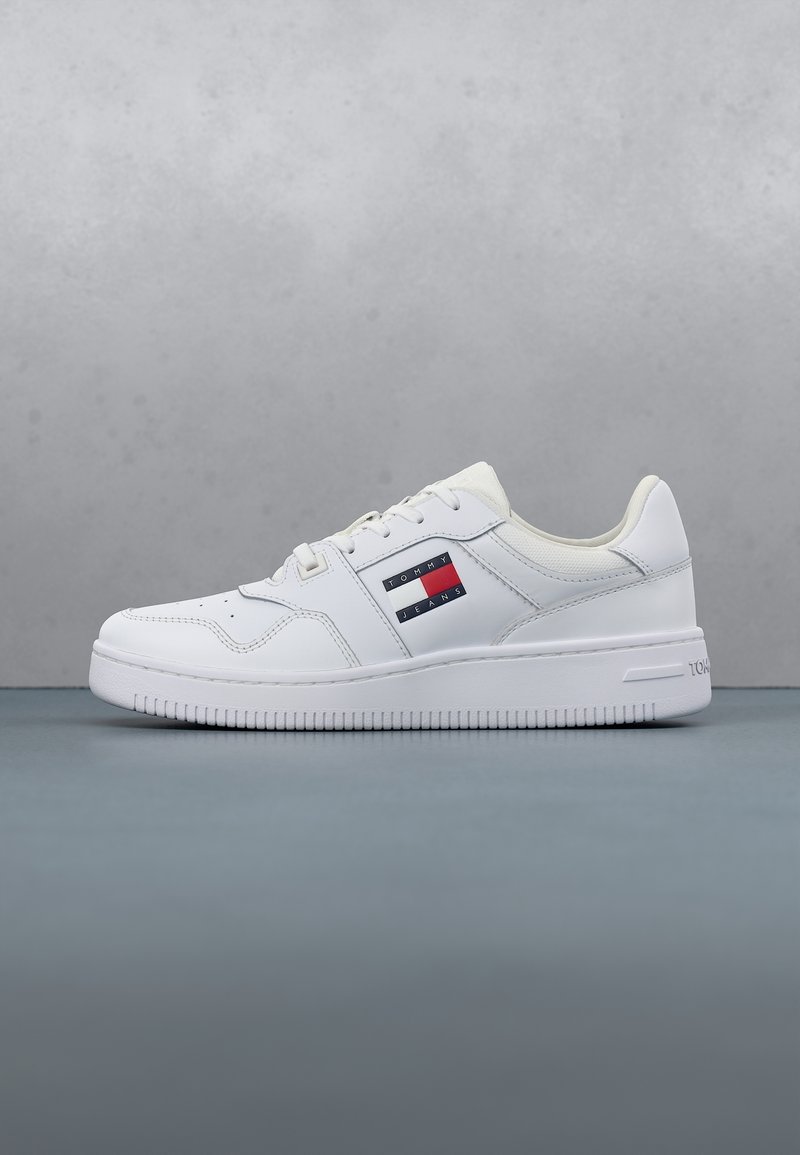 Baskets blanches basses avec fermeture à lacets, logo Tommy Jeans sur le côté et semelle texturée sur fond et surface gris.