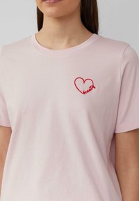 Lichtroze katoenen t-shirt met een ronde hals, met daarop een rode geborduurde hart met het woord "mooi" eronder.