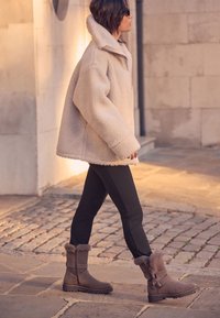 Veste en fausse fourrure beige, leggings noirs et bottines marron à accents en fourrure avec fermeture éclair latérale. La scène présente un pavé en pierre et un éclairage doux.