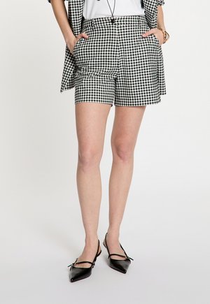 Mujer vistiendo shorts de vichy en blanco y negro con blazer a juego, top blanco, pulsera dorada y tacones slingback negros de punta afilada.