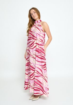 Ženska v brezrokavni maxi obleki z visokim ovratnikom, roza in rdečim vzorcem črt zebre ter žepi, stoji ob svetlem, enobarvnem ozadju.