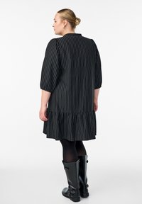 Nainen, jolla on vaaleat hiukset nutturalla, pukeutuneena mustaan raidalliseen mekkoon, jossa on pussitaskut, mustiin legginsseihin ja polvipitkiin mustiin saappaisiin, kääntyy vasemmalle.