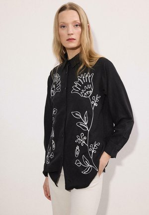 Femme aux longs cheveux blonds portant une chemise noire à boutons avec des motifs floraux blancs, debout devant un fond clair uni.