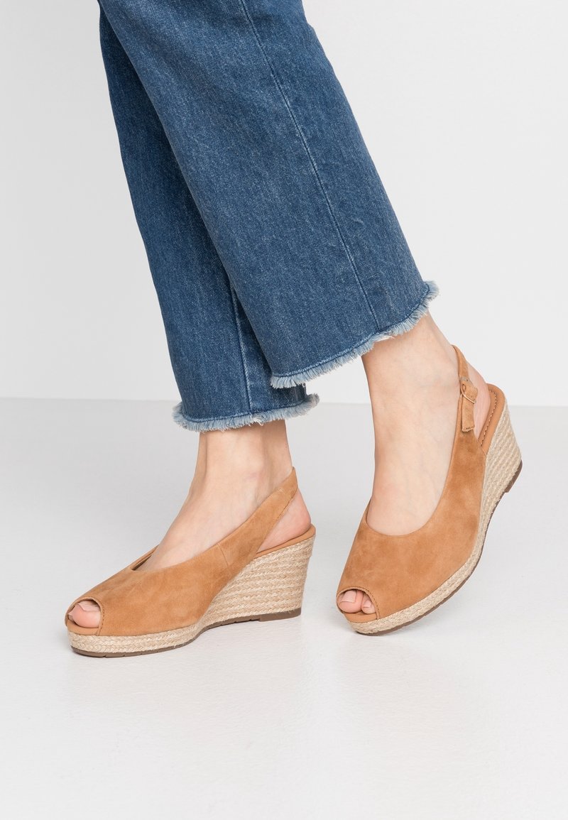 Pieds chaussés de sandales compensées en daim beige à bout ouvert avec brides autour de la cheville, portées avec un jean bleu coupé avec ourlet effiloché, debout sur un sol clair.