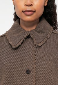 Vrouw die een bruin gestructureerd button-up jasje met een ruime kraag en rafelige randen draagt, zichtbaar van kin tot bovenste borst.