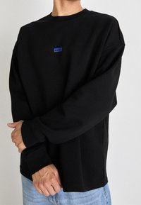 Sudadera negra con cuello redondo, mangas largas, puños de canalé y un pequeño logotipo azul etiquetado como "HUGO" en el pecho. Textura suave.
