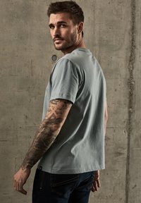 Světle modré tričko s crew neck výstřihem vyrobené z bavlny, s krátkými rukávy a volným střihem, v kombinaci s tmavými džínami a propracovanými tetováními na rukávech.