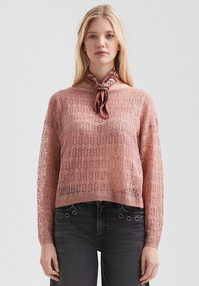 Blonde Frau in einem rosa, aus Spitze gestrickten Pullover, schwarzen Jeans mit Ösen-Details und einem mauvefarbenen gemusterten Halstuch, das vor einem schlichten Hintergrund steht.
