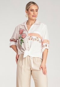 Biała i różowa koszula w paski z kwiatowym haftem, zapinana na guziki, związywanym przodem, podwiniętymi rękawami i luźnym krojem.